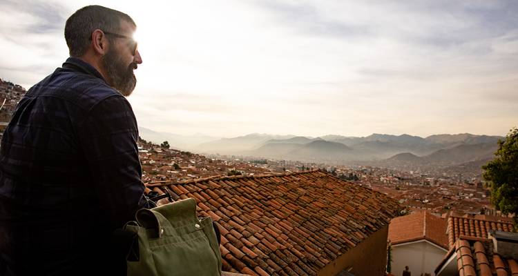 Un voyageur avec un sac à dos contemple les toits en terre cuite qui surplombent une vaste ville de montagne baignée dans la douce lumière du soir.
