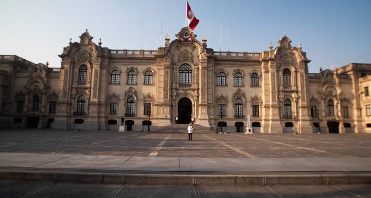 Guardia peruano de pie frente al ornado Palacio de Gobierno en la plaza principal de Lima bajo cielos despejados.