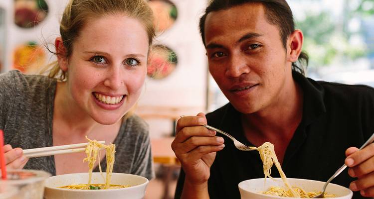 Deux convives souriants dégustant des bols de soupe de nouilles avec des baguettes et des cuillères.