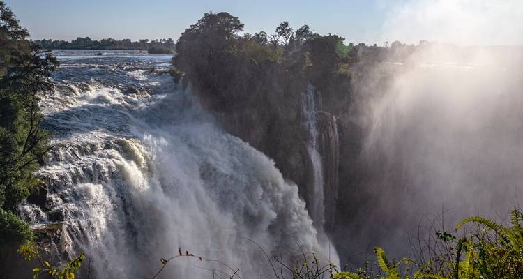 De puissantes chutes d'eau se déversent sur les chutes Victoria avec de la brume s'élevant dans l'air ensoleillé.