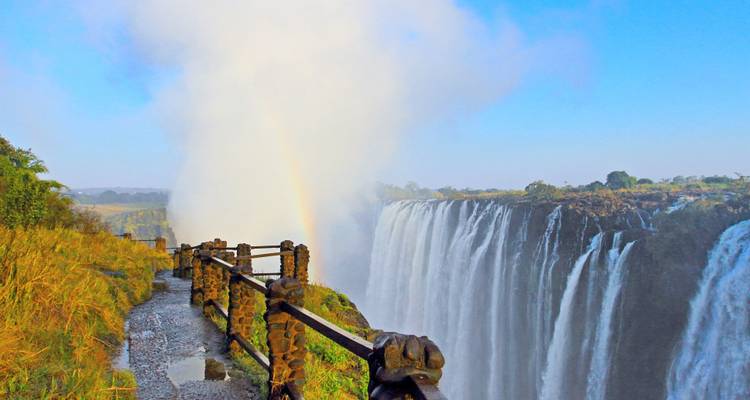 Rideau de brume des chutes Victoria plongeant dans une gorge avec un arc-en-ciel arqué au-dessus d'un sentier en corniche.