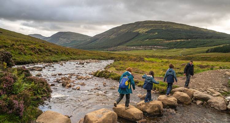 Une famille traversant de grandes pierres au-dessus d'un ruisseau par un jour nuageux dans une vallée des Highlands.