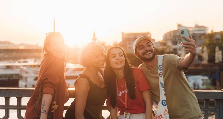 Quatre amis prenant un joyeux selfie sur un pont au coucher du soleil avec la ligne d'horizon d'Istanbul doucement éclairée derrière eux.