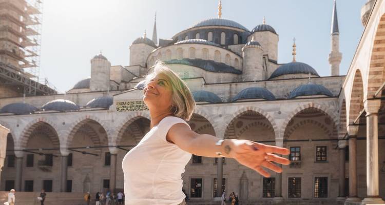 Femme souriante avec les bras tendus posant devant les dômes et les minarets de la Mosquée Bleue.