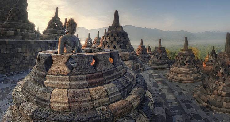 Zonneschijnlicht over stenen stupas en Boeddhabeelden in de tempel van Borobudur met mistige heuvels op de achtergrond.