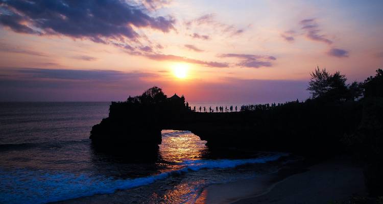 Dramática silueta del atardecer del templo marino Tanah Lot encima de un arco rocoso sobre olas que se estrellan.