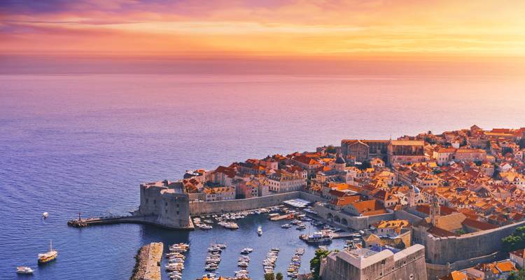 Vista aérea del atardecer de la ciudad antigua fortificada de Dubrovnik y su puerto brillando junto a las tranquilas aguas del Adriático.