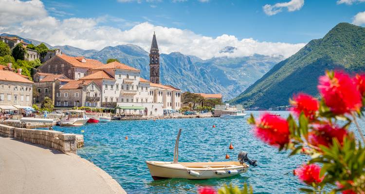 Encantador pueblo frente al mar de Perast con torre de iglesia de piedra, montañas y flores brillantes en primer plano.