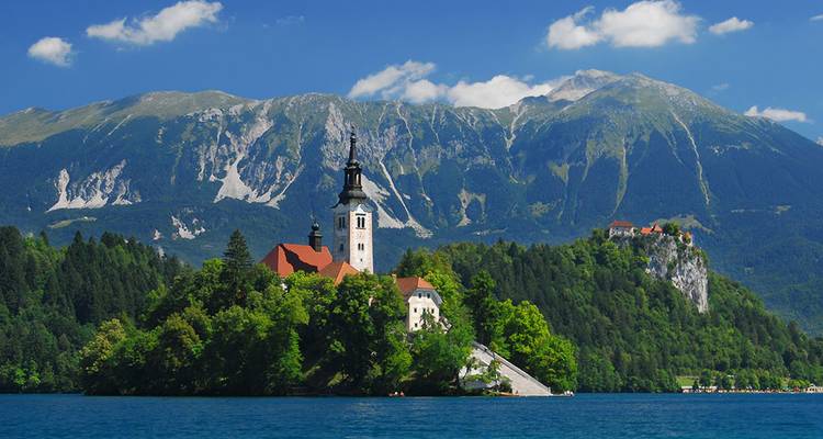 Église pittoresque sur une petite île boisée dans un lac d'un bleu cristallin avec des montagnes alpines spectaculaires s'élevant à l'arrière-plan sous un ciel ensoleillé.