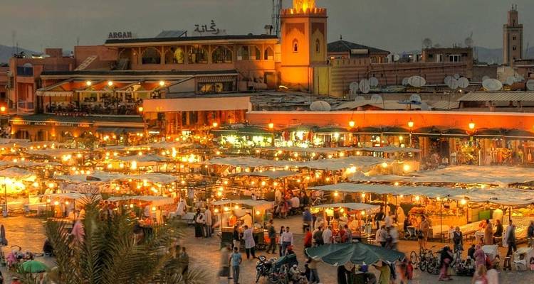 Vista nocturna de la plaza Jemaa el-Fnaa llena de puestos de comida brillantes y multitudes bajo el cielo del atardecer.