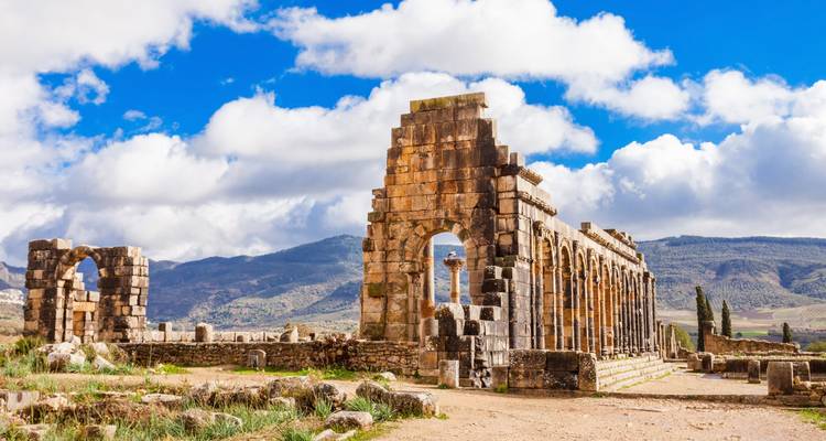 Arcos de piedra de las ruinas romanas en Volubilis con colinas ondulantes y nubes esponjosas al fondo.