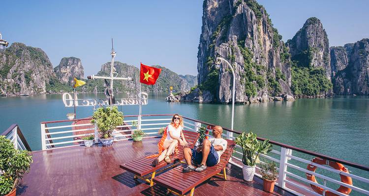 Un couple se détend sur des chaises longues d'un bateau de croisière, savourant un café avec les îles calcaires imposantes de la baie d'Halong qui les entourent sous un ciel bleu dégagé.