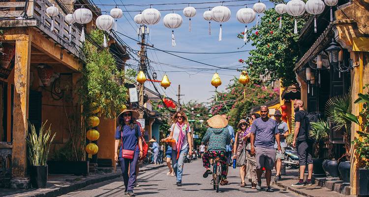 Les visiteurs se promènent et font du vélo dans une rue de la vieille ville de Hoi An bordée de lanternes, sous un soleil éclatant et des décorations festives.
