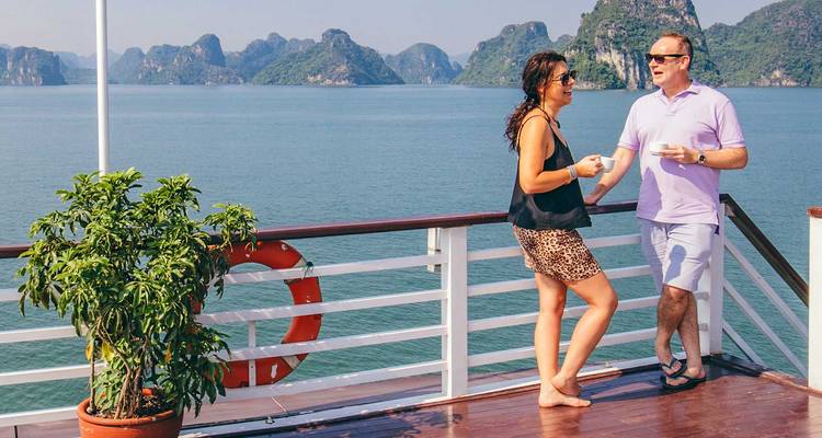 Una pareja disfruta de bebidas en una terraza de madera mientras contempla las tranquilas aguas color esmeralda y los picos de piedra caliza de la Bahía de Halong.