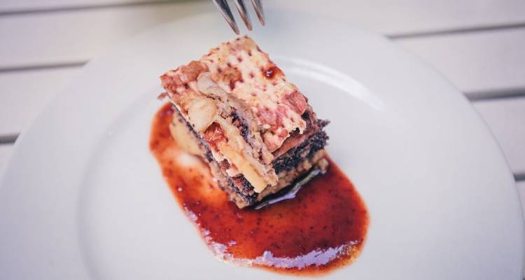 A plated slice of layered pastry dessert sits in a pool of red fruit sauce on a white dish.
