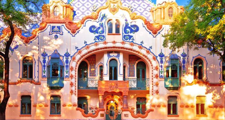 A colourful Art Nouveau façade adorned with blue floral motifs glows in warm sunlight behind leafy trees.