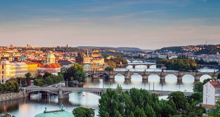 Panoramic view over Prague’s red roofs and multiple historic bridges spanning the Vltava River at sunset.
