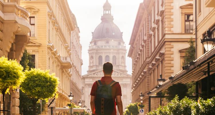 A backpacker walks toward Budapest’s St. Stephen’s Basilica, sunlight streaming between grand buildings.