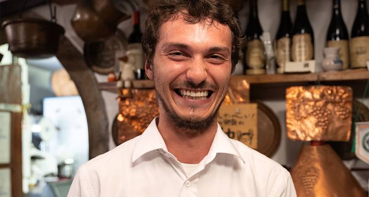 Portrait d'un serveur joyeux dans une trattoria italienne accueillante, avec des bouteilles et des casseroles en cuivre derrière lui.