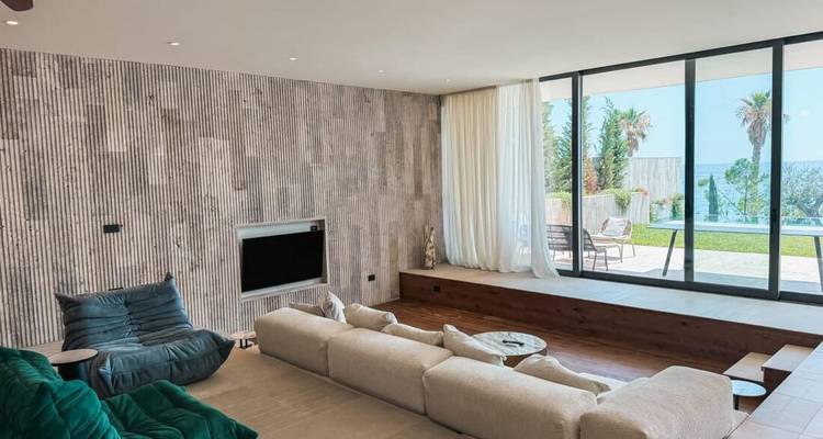 Modernes Wohnzimmer einer Küstenvilla mit großem Sofa, raumhohen Fenstern und Poolblick.