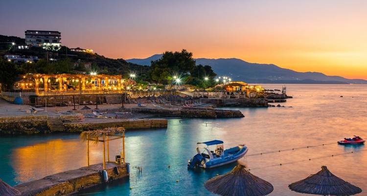 Küstendorf mit beleuchteten Restaurants an der Uferpromenade und ruhigem türkisfarbenem Wasser bei Sonnenuntergang.