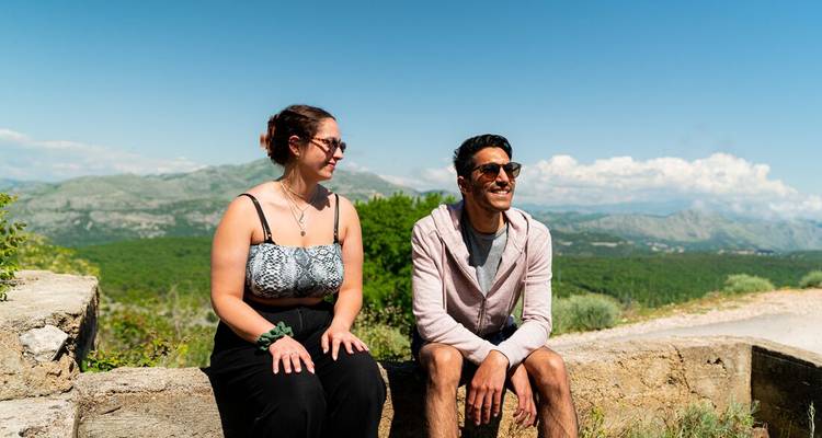 Junges Paar sitzt auf einer Steinmauer und genießt die Panoramablicke auf die Berge unter klarem Himmel.