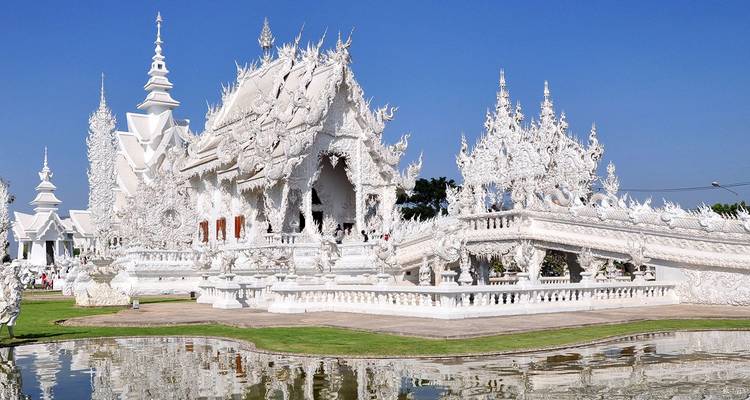 Schitterend witte Wat Rong Khun tempel met ingewikkelde uitsnijdingen weerspiegeld in een vijver onder een diepblauwe hemel.