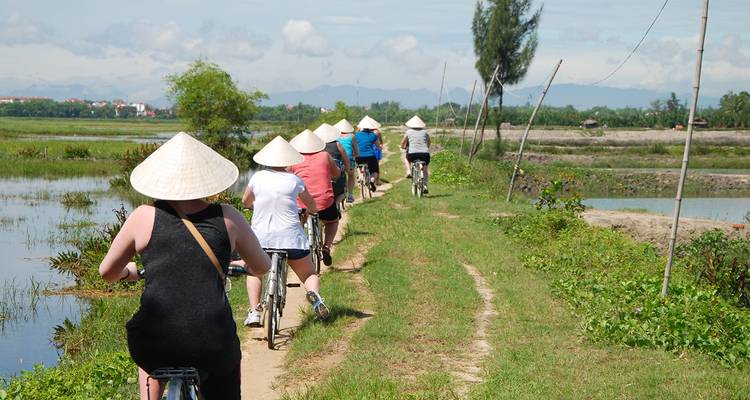 Rangée de cyclistes portant des chapeaux coniques qui roulent le long d'un sentier de terre étroit à côté de rizières et de montagnes lointaines