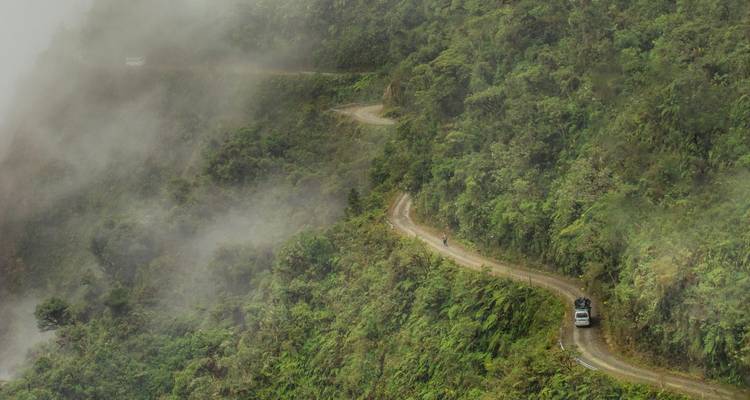 Carretera de montaña sinuosa que desaparece en la niebla con un vehículo solitario navegando por la ladera exuberante del acantilado