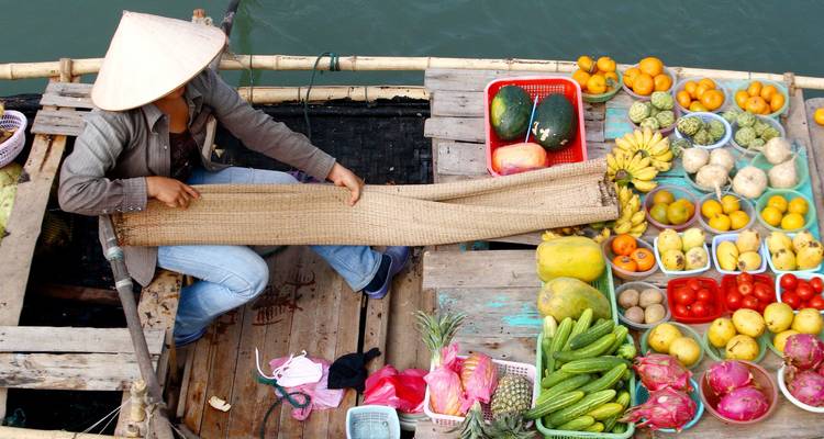 Marktverkoper op de drijvende markt met een punthoed verkoopt een verscheidenheid aan kleurrijke groenten en fruit vanaf een houten bootje.