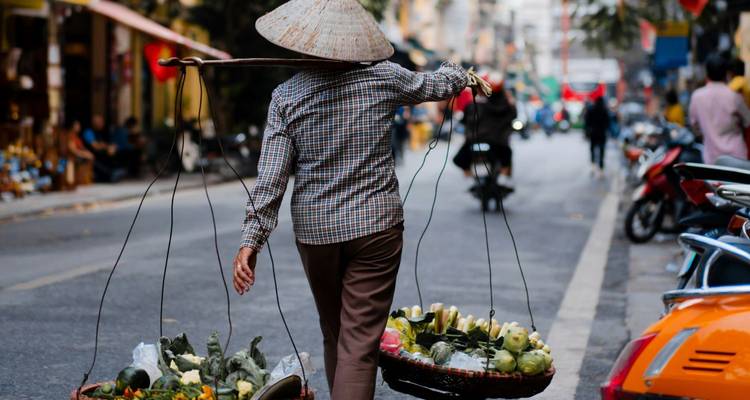 Een straatvendelaar balanceert twee productkandelen op een draagstok op zijn schouder terwijl hij door een drukke stadsstraat loopt.