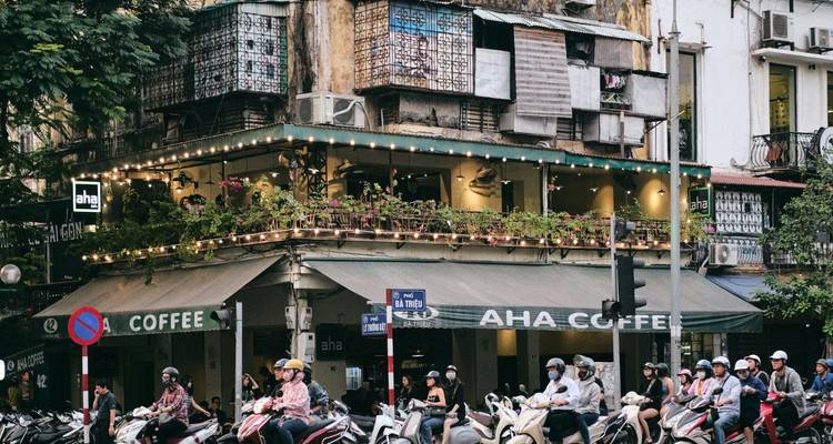 Belebte Hanoi-Straßenecke mit einem Kolonialgebäude, das ein Cafe beherbergt, und Motorradströmen, die vorbeifließen