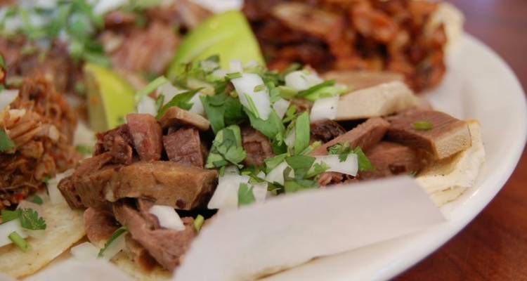 Close-up plate of tacos topped with tender meat, onions, cilantro and lime wedges.