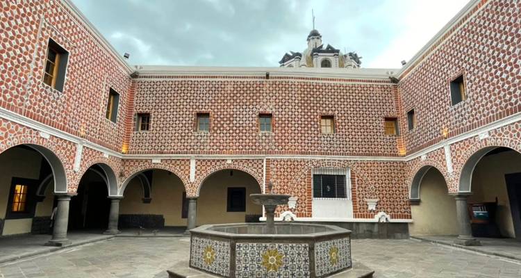 Peaceful colonial courtyard with patterned red-and-white tiles, arches and a central fountain under cloudy skies.