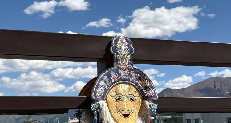 Masque tibétain décoratif exposé contre une balustrade avec un ciel bleu et des montagnes à l'arrière-plan.