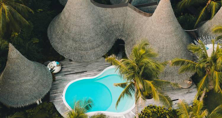 Vue aérienne d'une station balnéaire de charme aux toits de chaume arrondis entourant une piscine turquoise parmi les palmiers
