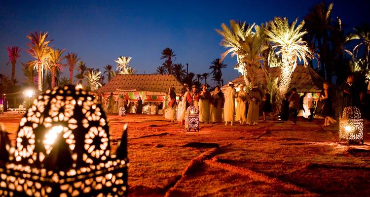 Nighttime desert camp festival with lanterns, palm trees and traditional dancing under dark sky.
