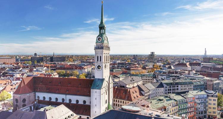 Vista panorámica de la ciudad con una alta torre de iglesia elevándose sobre edificios con tejados rojos bajo un cielo azul despejado.