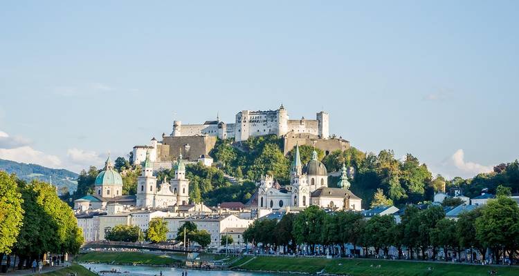 Ciudad europea junto al río con una fortaleza en la cima de la colina e iglesias con cúpulas rodeadas de árboles verdes.