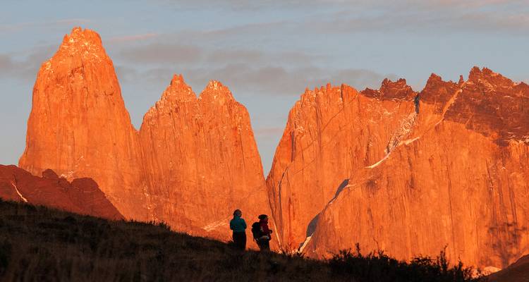 Twee wandelaars staan afgetekend tegen de vurige granieten torens van Torres del Paine die bij zonsondergang gloeien.