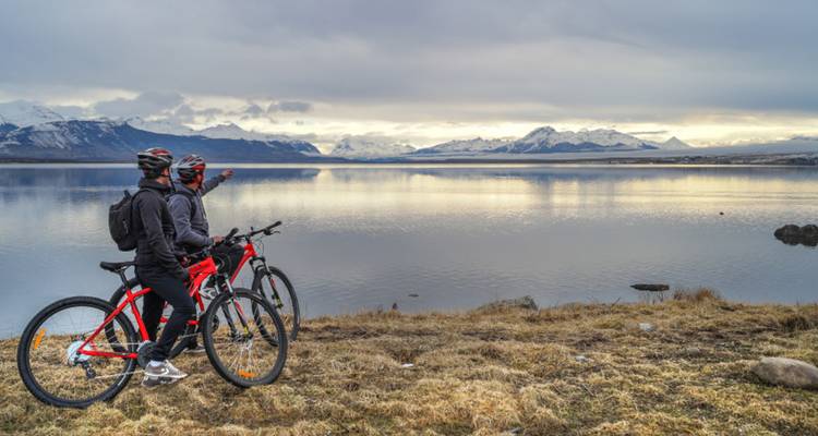 Twee fietsers maken een pauze op een grassy oever met uitzicht op een rustig Patagonisch meer en sneeuwbedekte bergen in de verte.