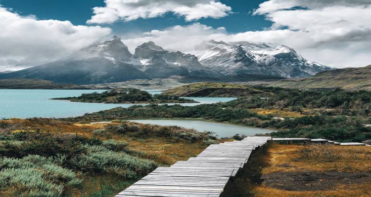 Een houten boardwalk leidt door weelderige struiken naar melkwitte turkooizen meren en gehavende sneeuwbedekte pieken onder dramatische wolken.