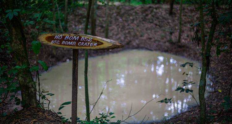 Un cratère de bombe boueux dans la jungle avec un panneau en bois indiquant « Cratère de bombe B52 ».