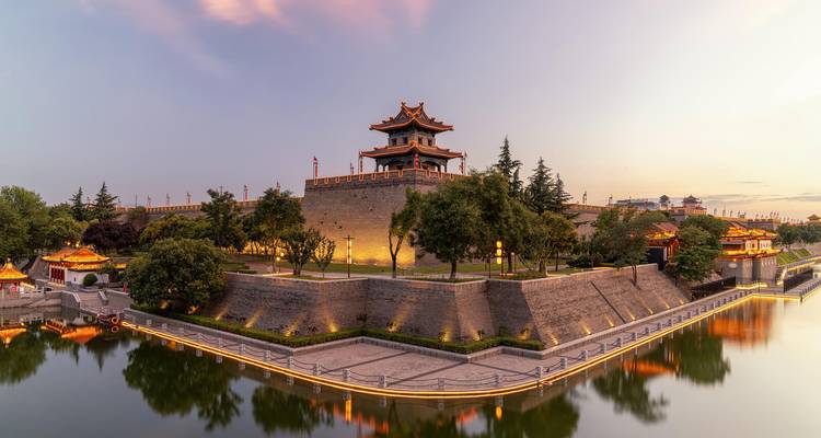 Panoramablick auf die antike Stadtmauer von Xi'an und den Wachturm, die bei Einbruch der Dunkelheit in warmem Licht erstrahlen und sich im umliegenden Graben spiegeln.
