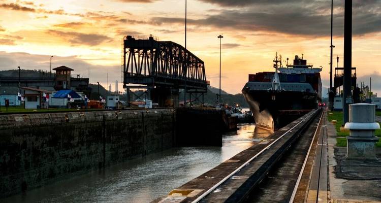 Un cargo traverse un sas du Canal de Panama au coucher du soleil, avec un pont et des tours de contrôle en silhouette.