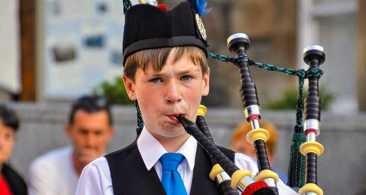 Ein junger Junge in traditioneller schottischer Kleidung spielt Dudelsack und konzentriert sich auf seine Aufführung, während verschwommene Zuschauer im Hintergrund zu sehen sind.