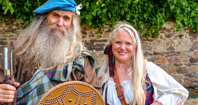Zwei Erwachsene in traditioneller schottischer Clan-Kleidung lächeln in die Kamera vor einer Steinmauer und Grünflächen.