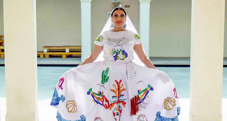 Woman in ornate traditional Mexican dress poses proudly in a bright colonnade.
