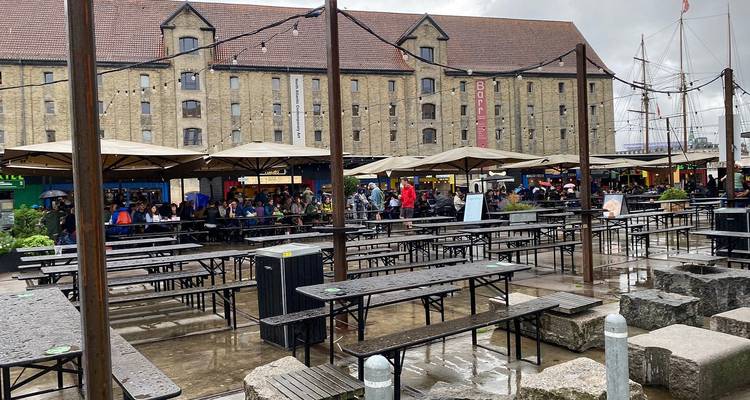 Marché alimentaire extérieur avec des bancs mouillés, des parapluies et des personnes se protégeant d'une légère pluie dans une cour d'entrepôt réaménagée.
