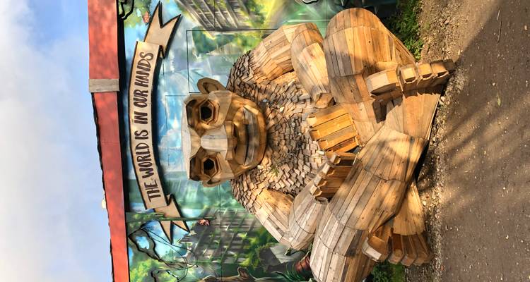Grande sculpture de troll en bois avec une bannière portant l'inscription « LE MONDE EST ENTRE NOS MAINS » devant une fresque murale colorée.